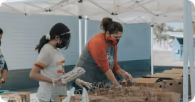 Two people wearing face masks sorting and packing items under an outdoor tent.