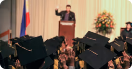 Audience of graduates in caps raising hands toward a speaker at a podium on stage with floral arrangement and flag backdrop.