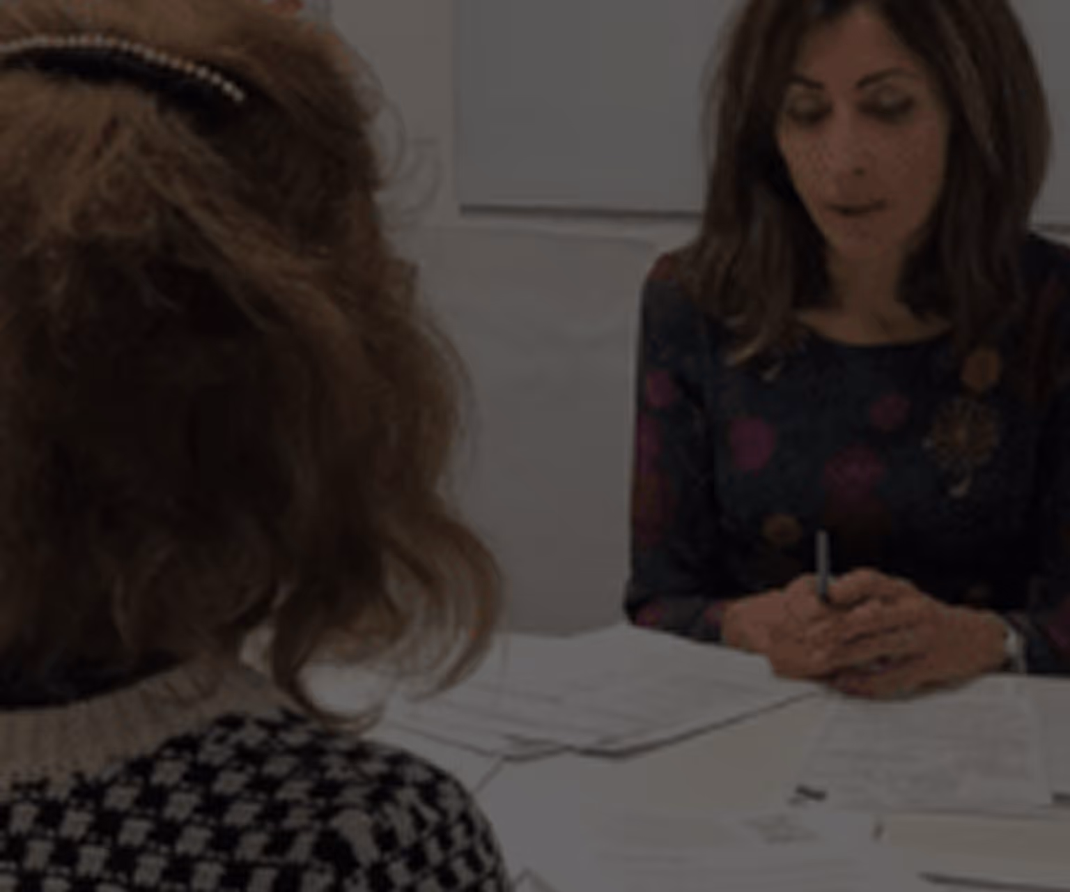 Two women sitting at a table covered with papers, one woman is facing the camera and speaking while the other has her back to the camera.