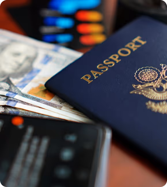 Close-up of a navy blue passport on top of US dollar bills with a blurred mobile phone and colored pens in the background.