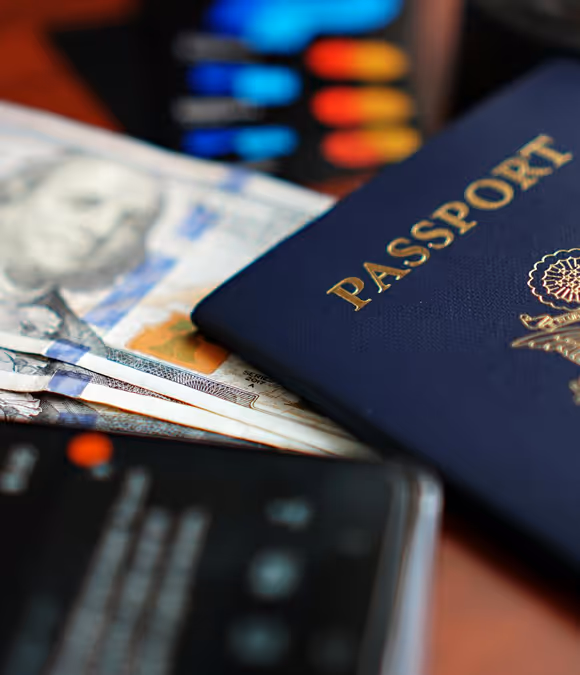 Close-up of a blue passport resting on US hundred-dollar bills with a blurred phone and colorful streaming device in the background.