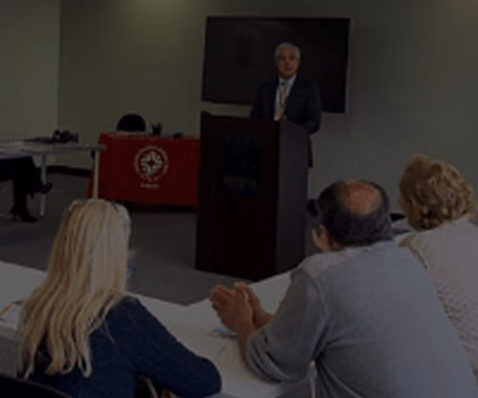 Man in a suit speaking at a podium in a conference room with three seated attendees listening.