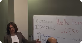 Woman sitting and gesturing next to a whiteboard with handwritten notes about checking savings accounts and Wells Fargo.