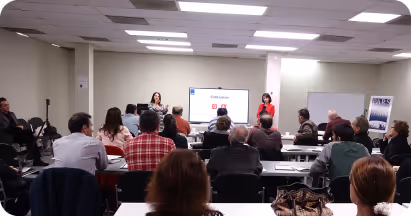 People seated in a classroom facing two presenters and a screen displaying a presentation in a conference room.