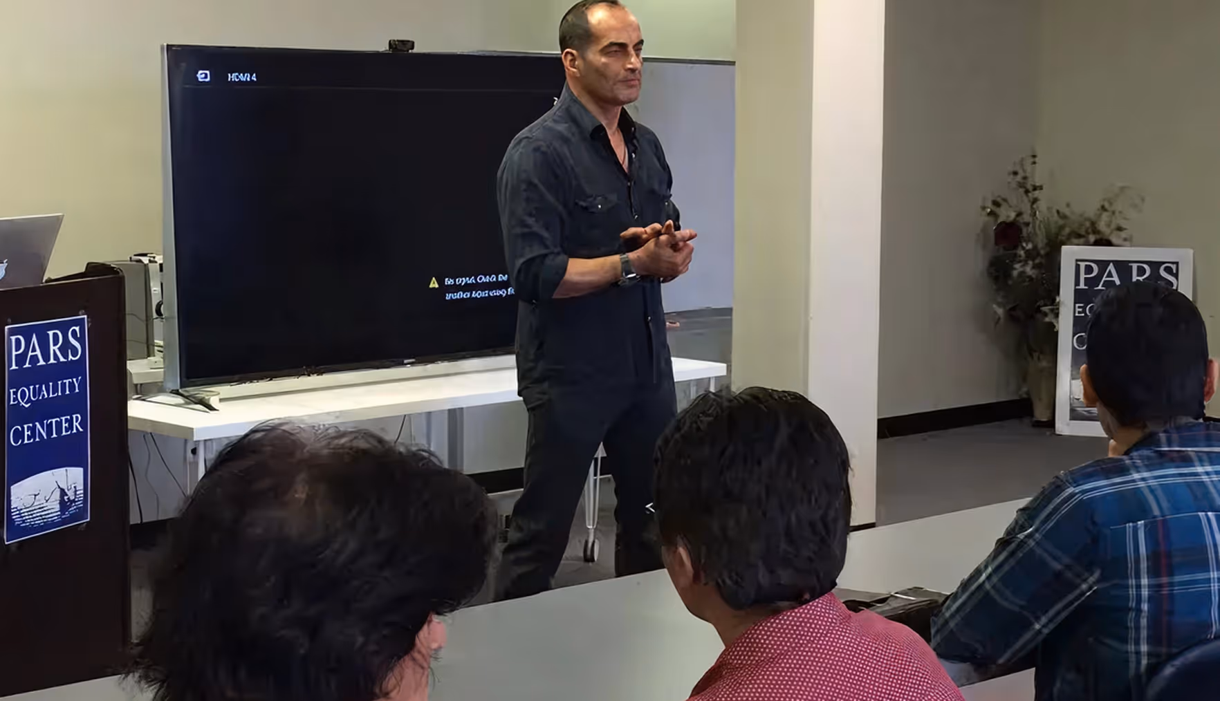 Man standing and speaking in front of a large screen in a meeting room with seated attendees.
