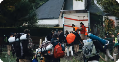 Group of hikers carrying backpacks and camping gear walking towards a white truck in a wooded area.