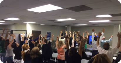 Group of people standing in a classroom raising their hands during an activity.