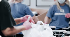 Volunteers wearing masks and gloves packing food items into white plastic bags indoors.