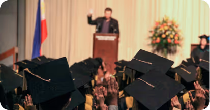 Graduates in caps raising hands while a speaker addresses them from a podium on stage.