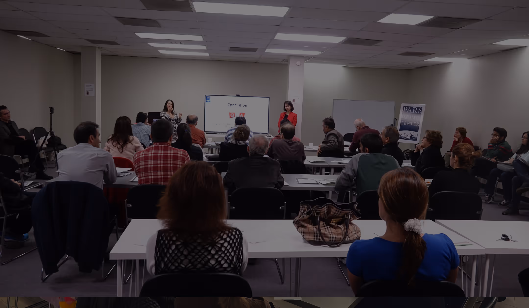 Audience seated in a conference room watching a presentation with a screen displaying 'Conclusion' and two presenters standing at the front.