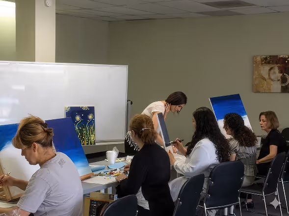 Group of women painting on canvases seated at tables in a classroom with a whiteboard.