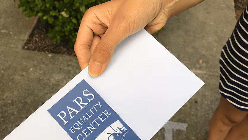 Person holding an envelope with a blue label reading Pars Equality Center outdoors.