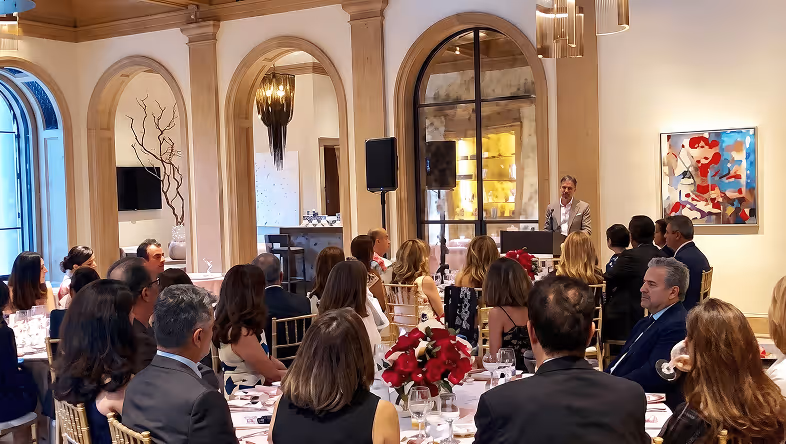 Audience seated at round tables listening to a speaker standing at a podium in a banquet hall.
