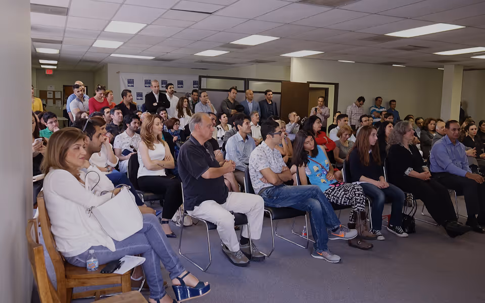 A diverse group of people seated and standing in a room attentively listening to a speaker.