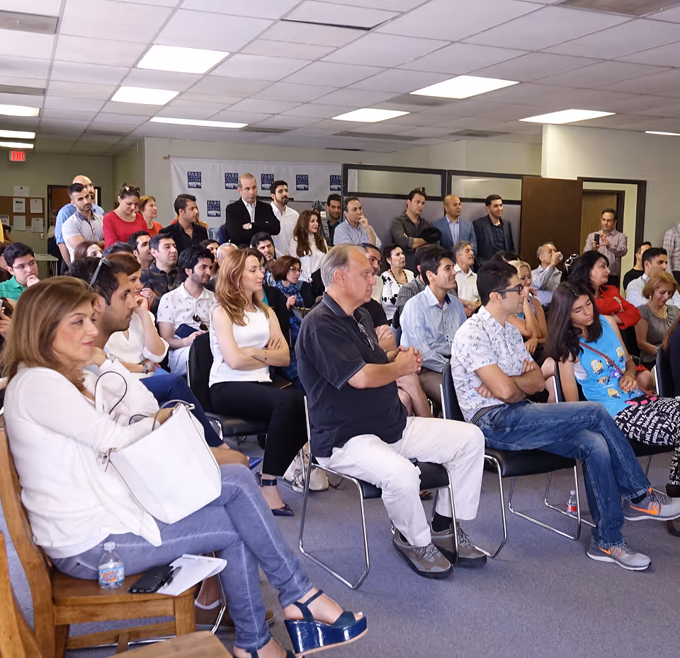 Audience of seated and standing people listening attentively at an indoor event in a conference room.