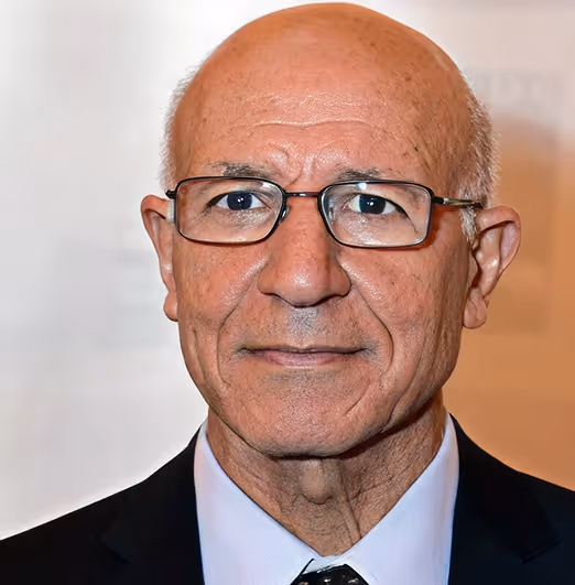 Headshot of an elderly man with glasses wearing a suit and tie against a plain background.