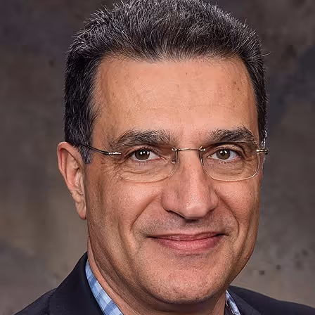 Smiling middle-aged man with glasses and short dark hair wearing a dark jacket and blue check shirt.