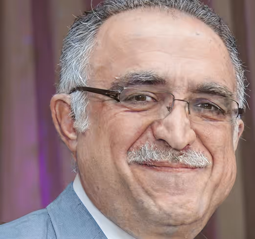 Close-up of a smiling elderly man wearing glasses and a light blue suit jacket.