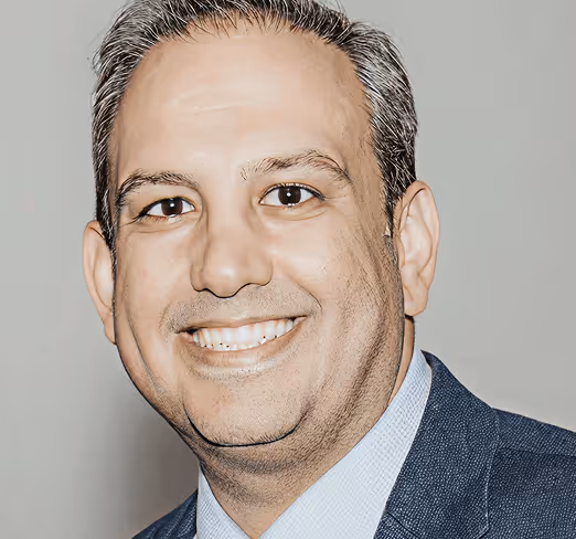 Smiling man with greying hair wearing a blue suit jacket and light blue shirt against a plain background.