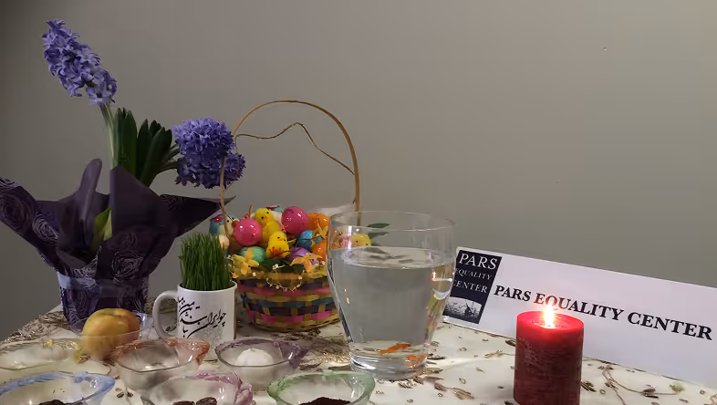 Table with purple flowers, grass, Easter basket, fishbowl, red candle, and Pars Equality Center sign.