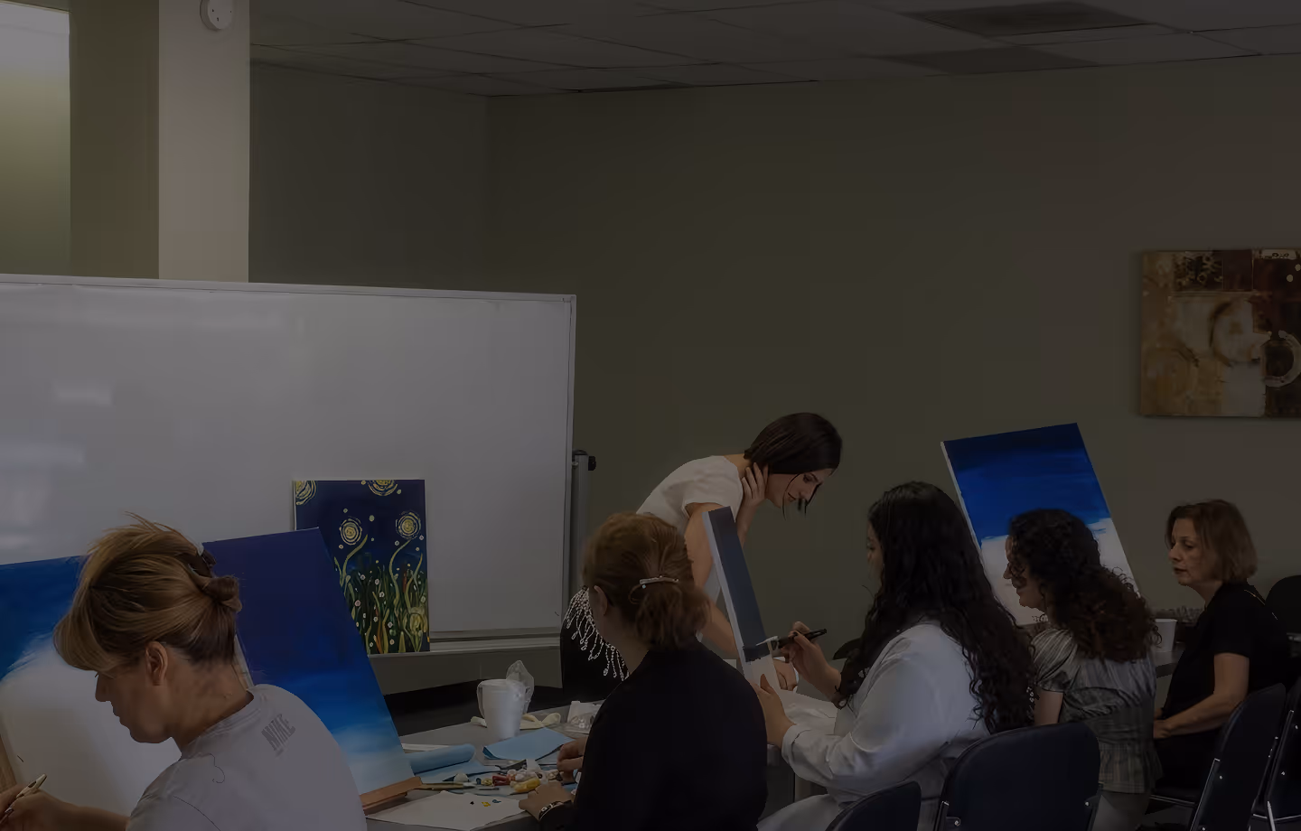 Group of women seated at tables painting on canvases in a classroom setting.
