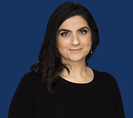 Woman with dark hair and earrings wearing a black top against a blue backdrop.