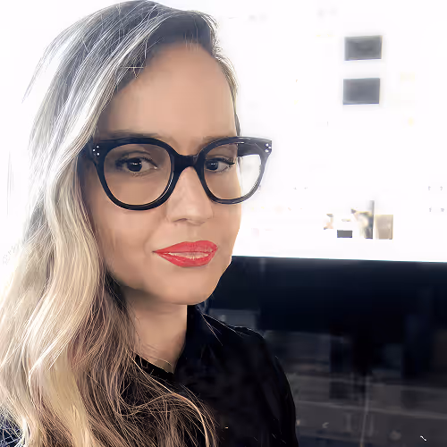 Woman with long blonde hair, black cat-eye glasses, and red lipstick smiling slightly indoors.