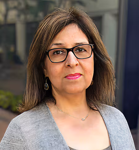 Portrait of a woman with glasses, pink lipstick, and shoulder-length brown hair wearing a gray top outdoors.