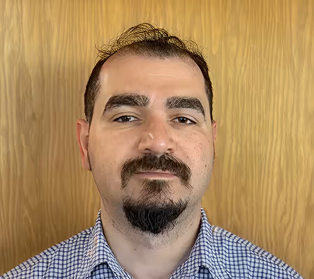 Man with dark hair, beard, and mustache wearing a checkered shirt against a wood panel background.