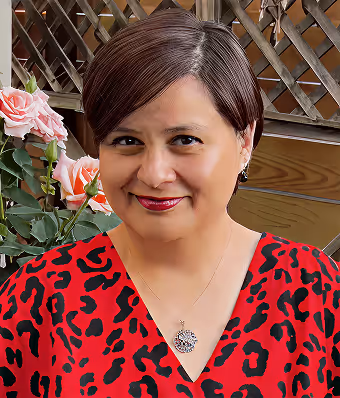 Smiling woman with short hair in a red and black patterned top, wearing a pendant necklace indoors.
