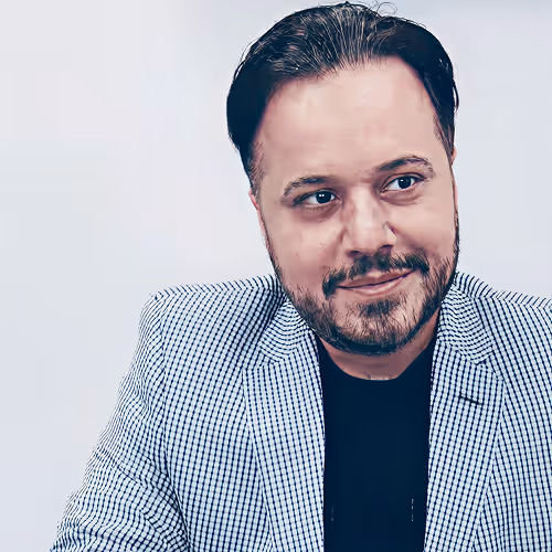 Man with a beard in a checkered blazer and black shirt, looking slightly to his right with a smile.
