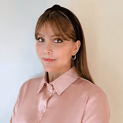 Smiling woman with light brown hair, black headband, hoop earrings, and pink button-up shirt.