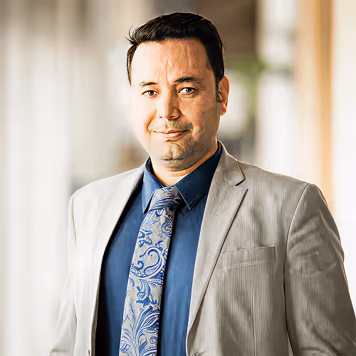 Man in beige suit jacket and blue shirt with patterned tie standing in a softly lit hallway.