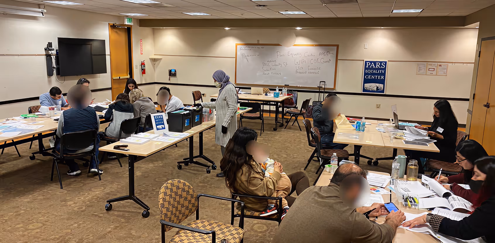 Several people seated and working at tables with papers and laptops in a room labeled Pars Equality Center.