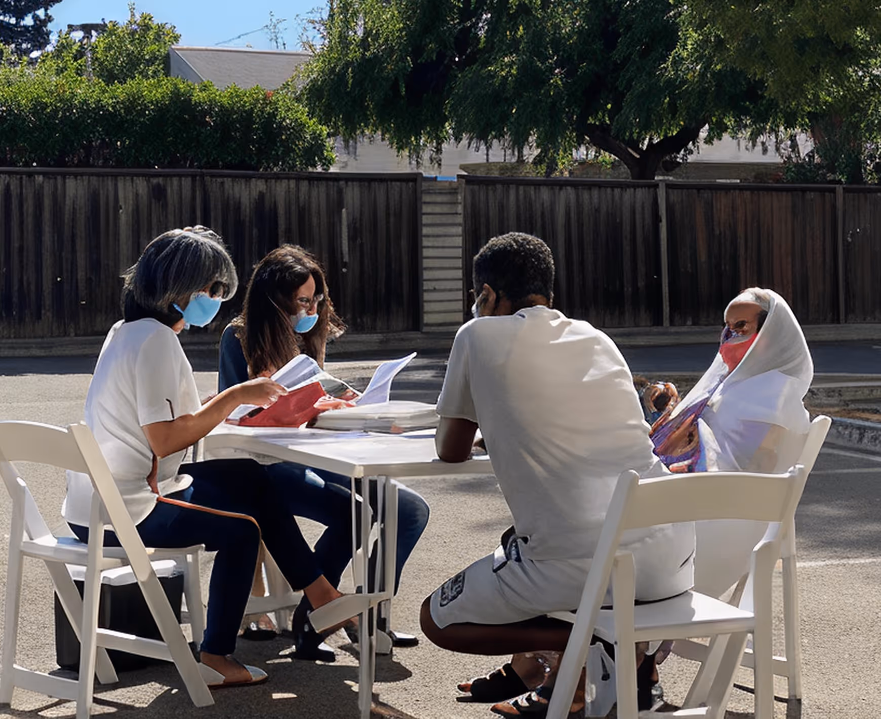 Four people wearing masks sitting outdoors around a table reading papers and documents.