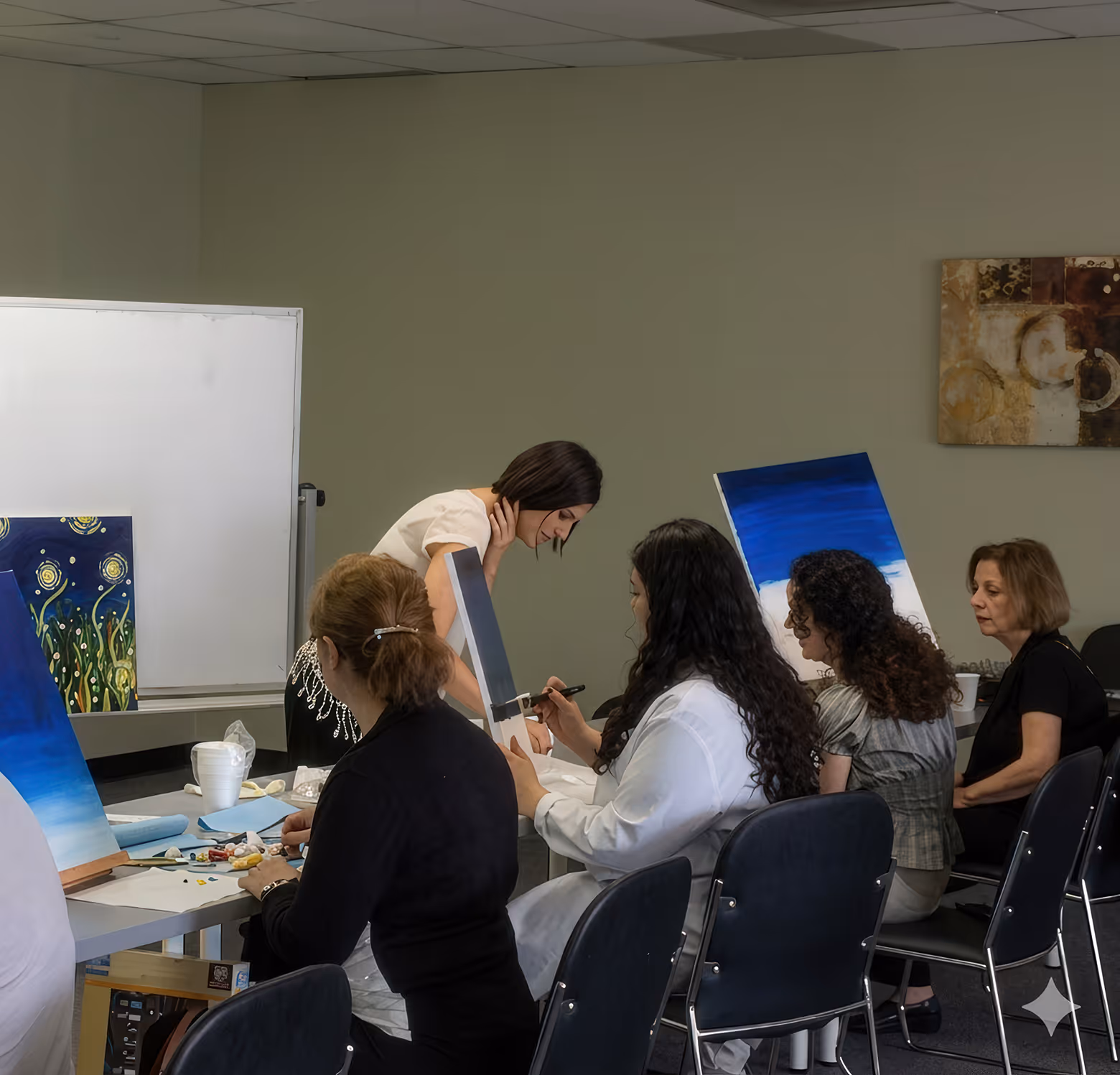 Group of women seated around a table painting on canvases in a workshop setting.