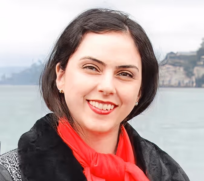 Smiling woman with dark hair, wearing a red scarf and black coat, outdoors by water with buildings in background.