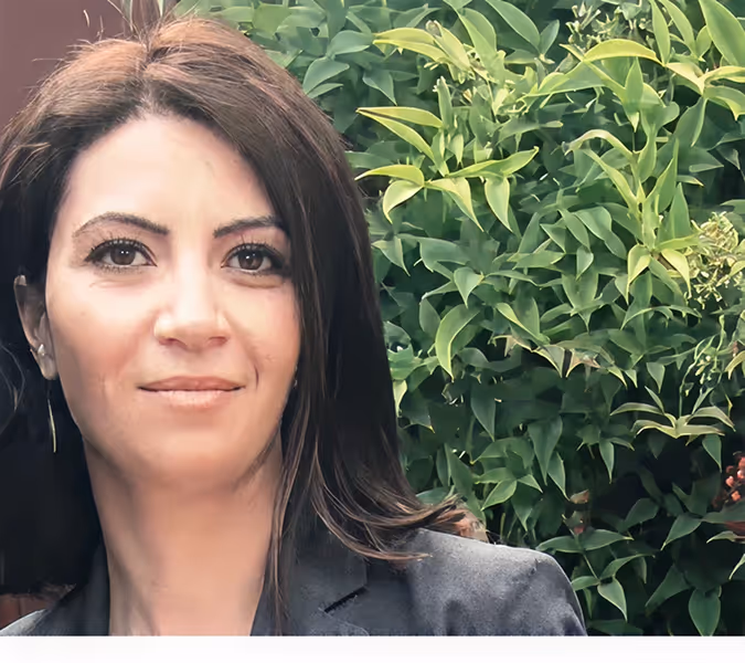 Woman with dark hair wearing a dark blazer, standing outdoors in front of green leafy bushes.