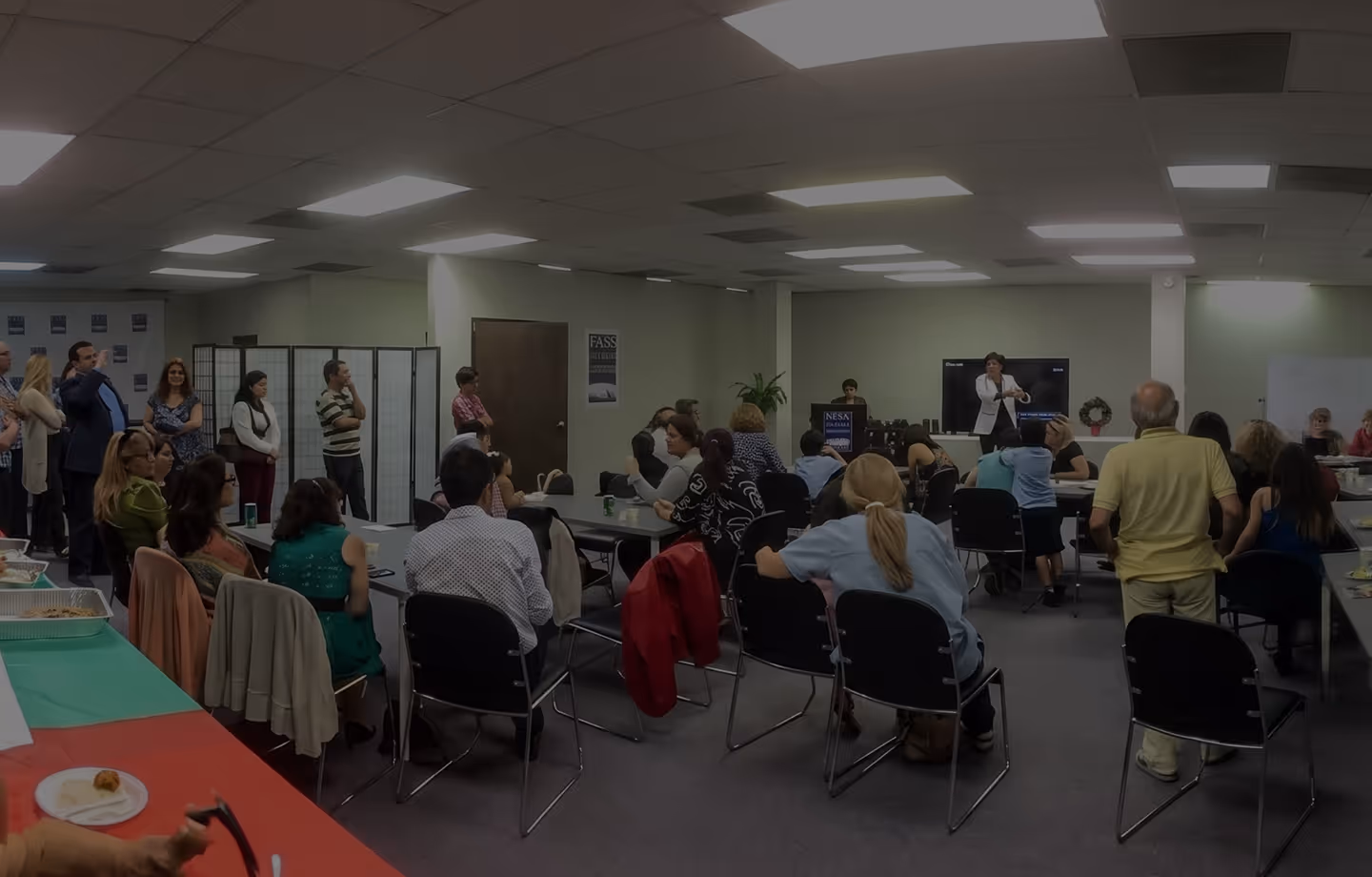 People seated and standing in a room attending a presentation with a speaker at front near a screen.