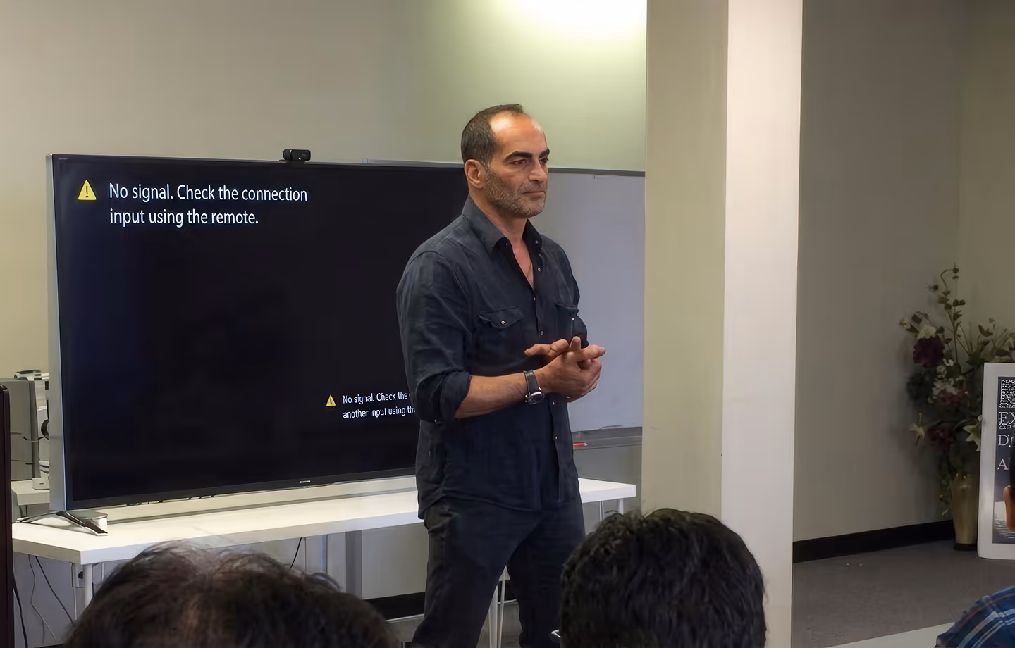 Man in dark shirt standing and speaking in front of a large screen showing no signal message.