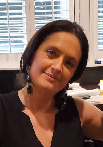 Woman with dark hair wearing earrings and a black top, posing indoors near window blinds.