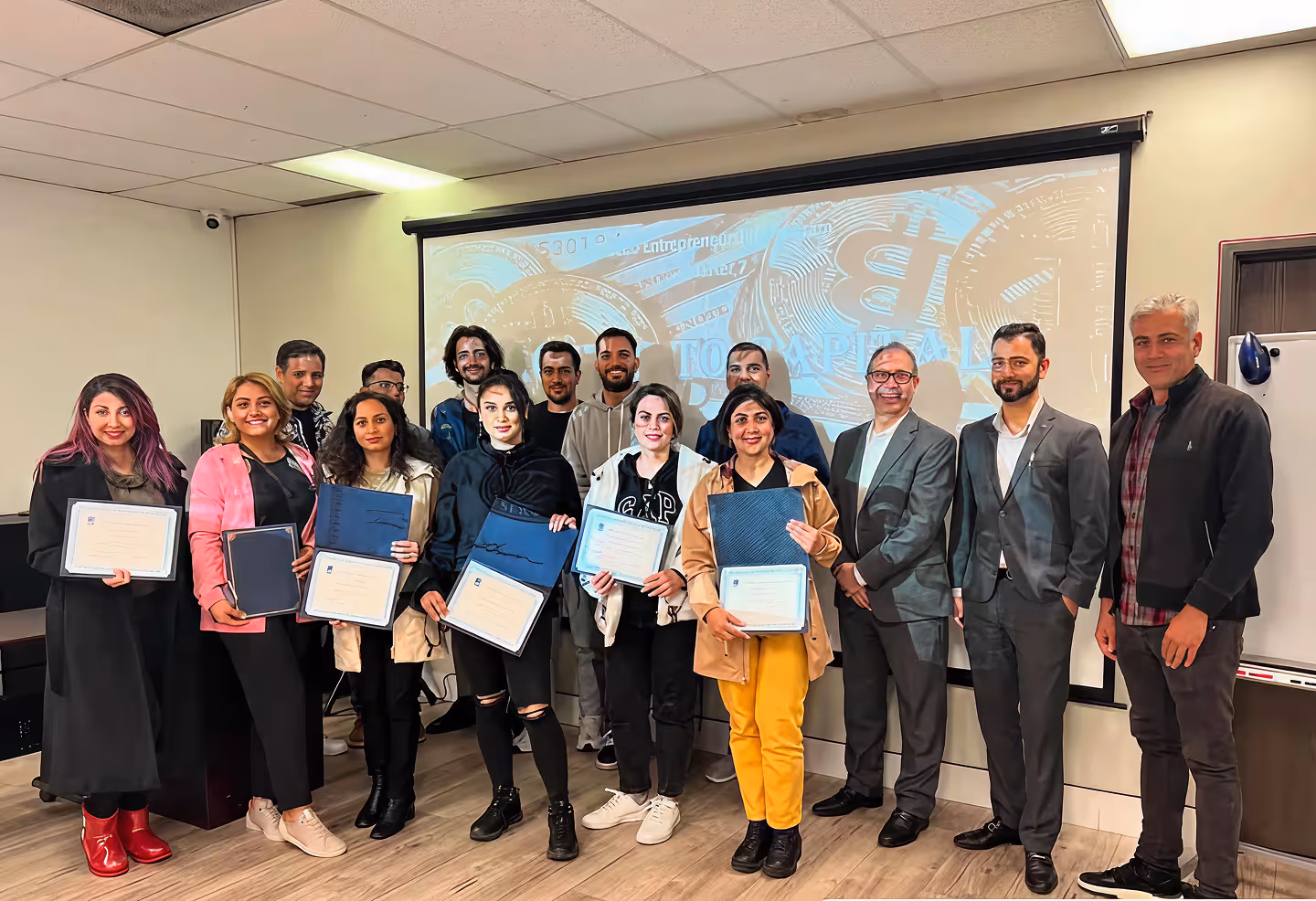 Group of men and women standing indoors, some holding certificates, with a projection screen behind them.