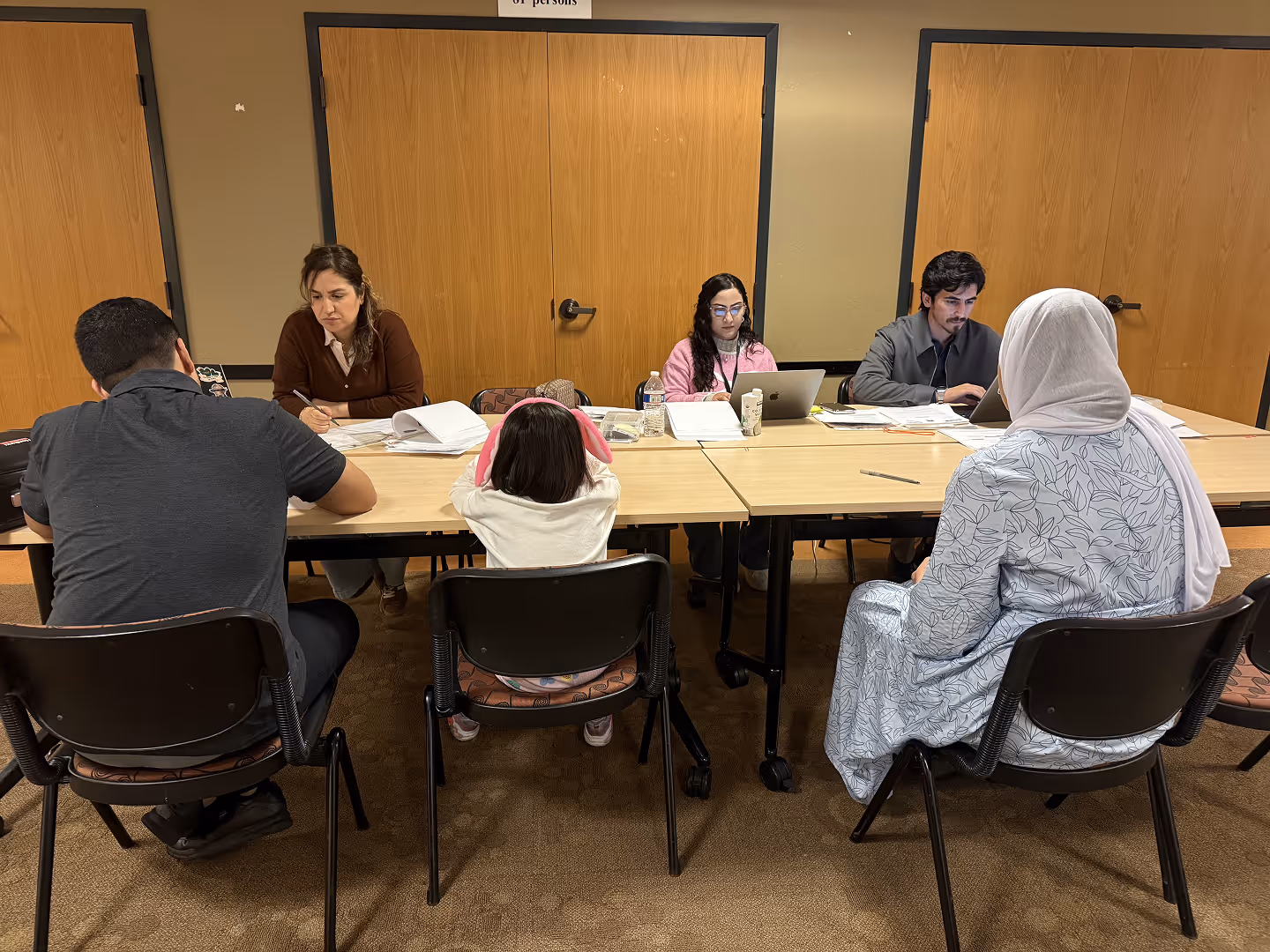 Four people sitting at a table with papers and laptops, and three people seated opposite them in a room.