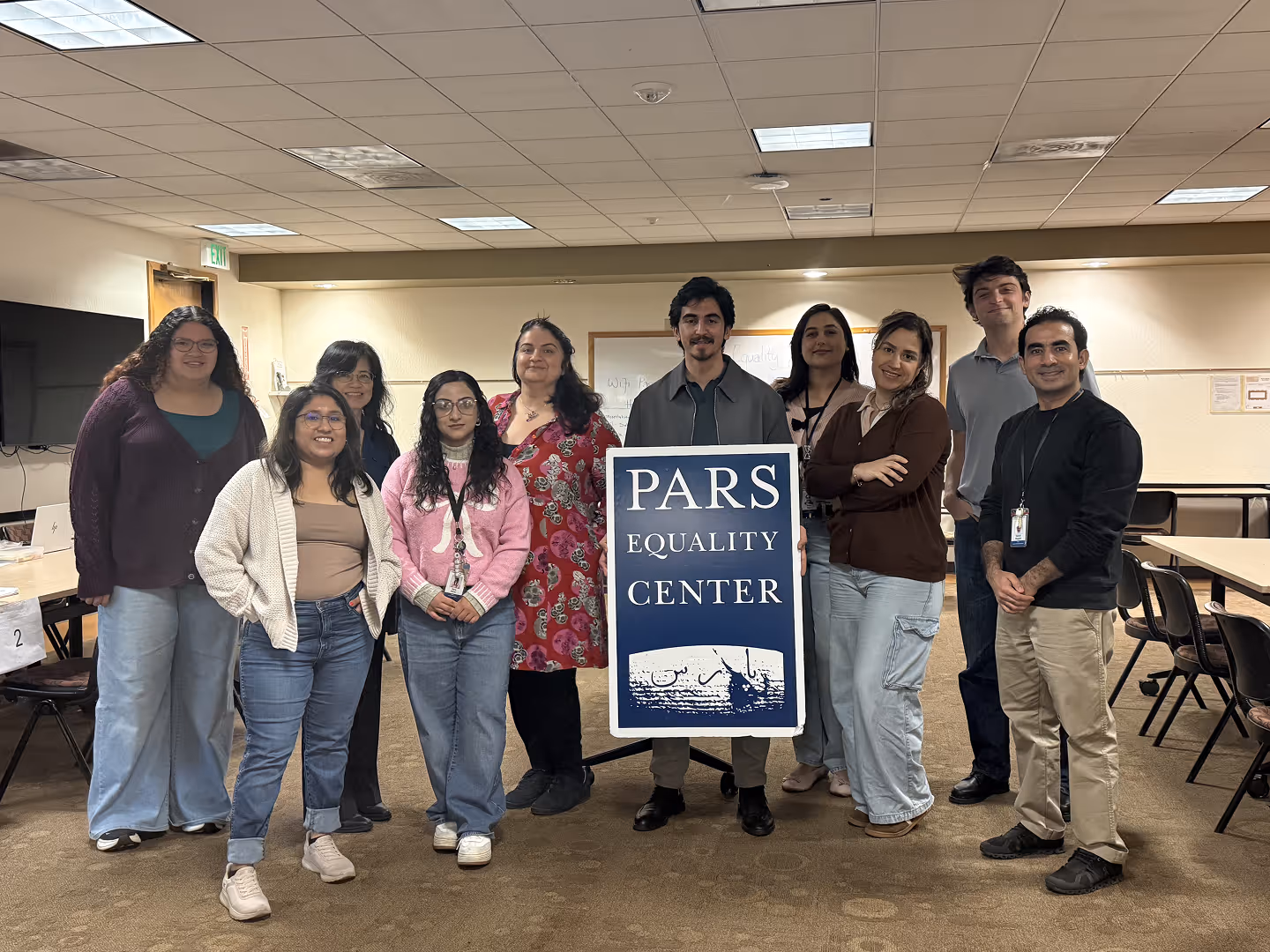 Group of ten smiling people indoors holding a Pars Equality Center sign in a meeting room.