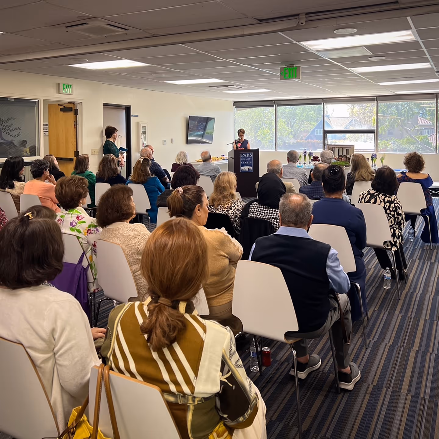 Woman at podium giving speech to seated audience in a bright meeting room with large windows.