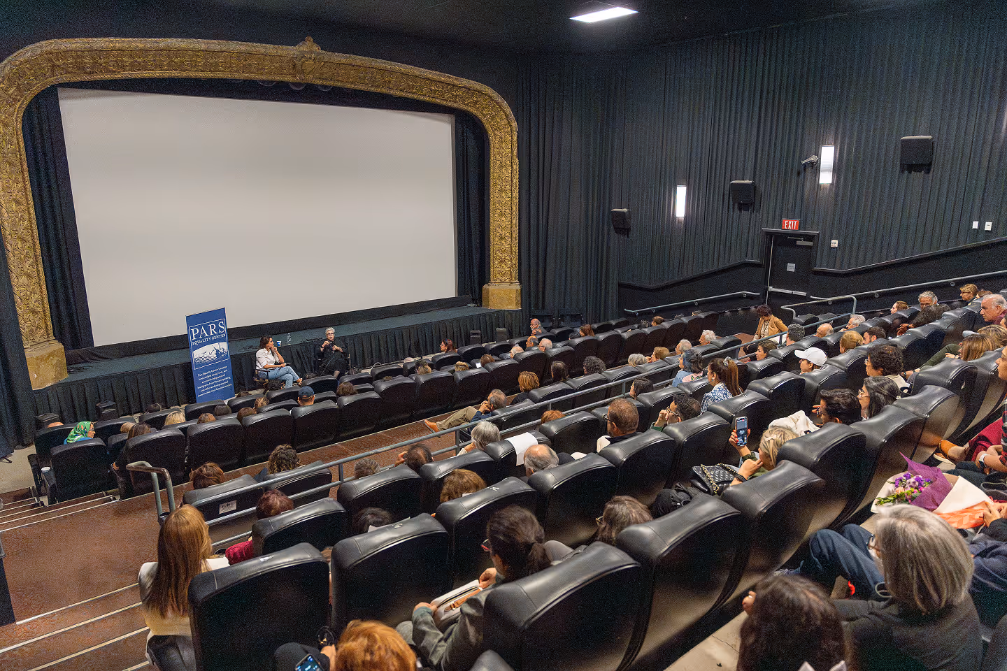 Audience seated in theater watching two people on stage speaking in front of a large blank screen.