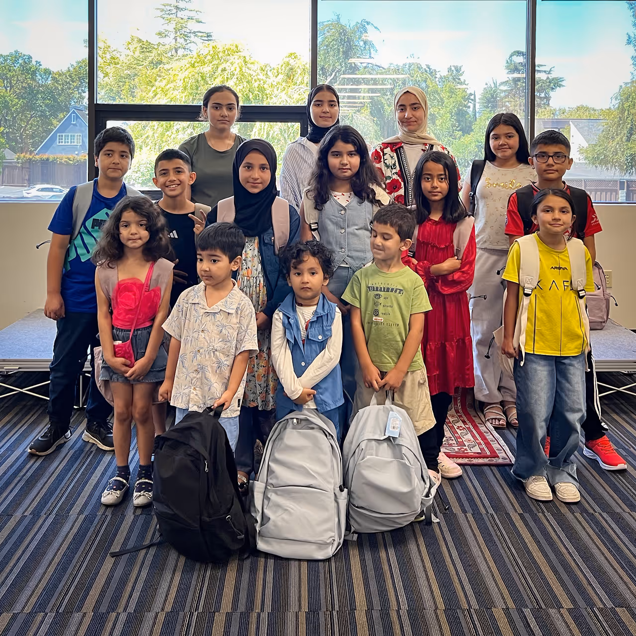 Group of children and teens posing indoors with backpacks near large windows showing trees outside.