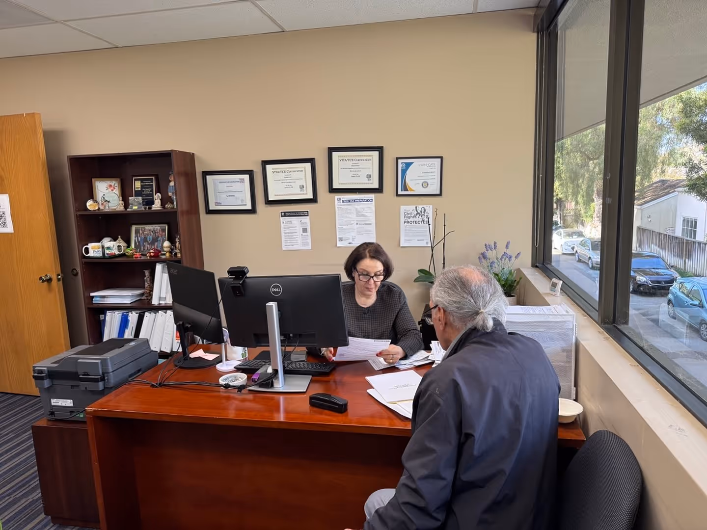 Office scene with a woman reviewing papers and an older man seated across her desk with a window view.