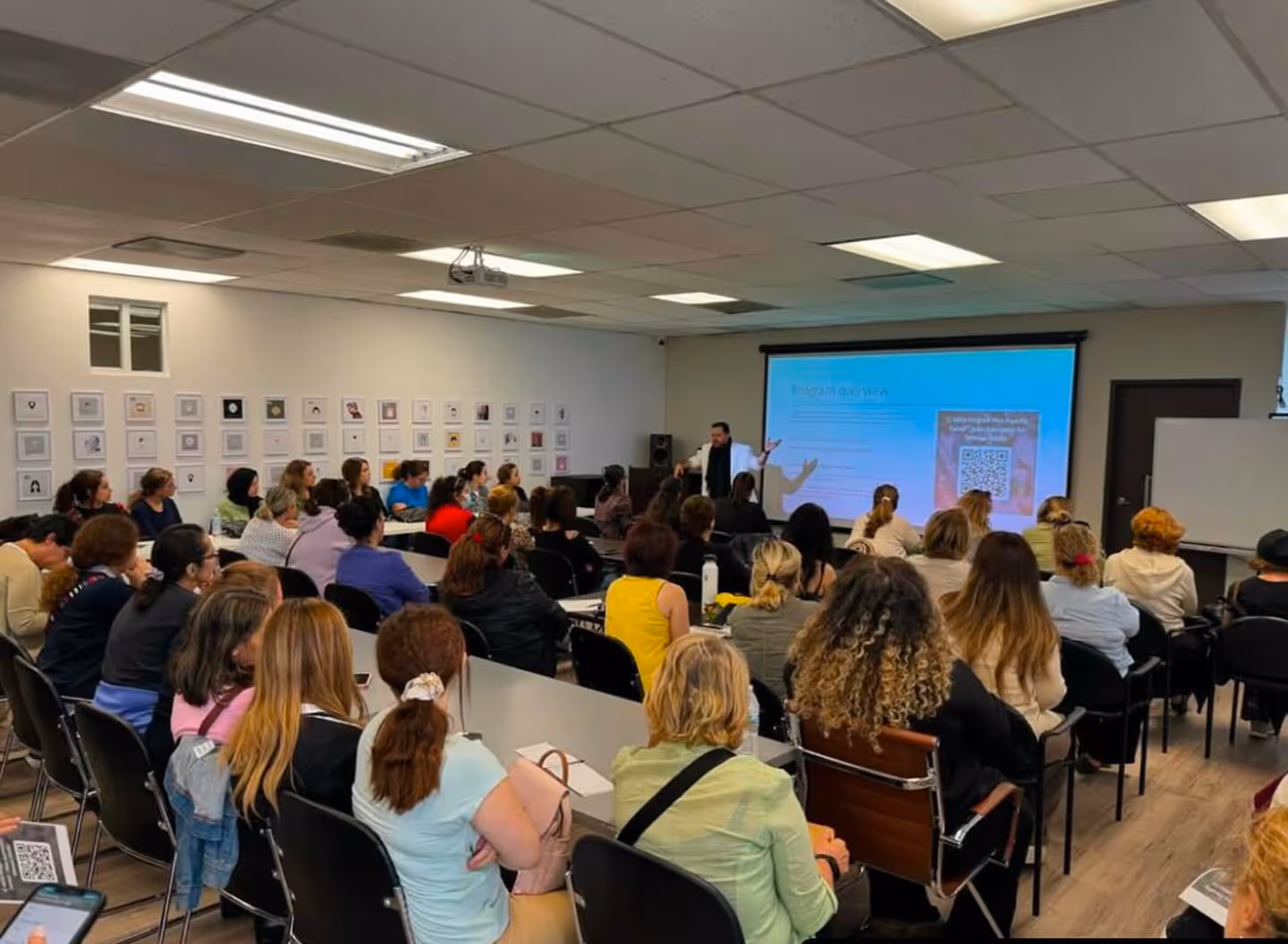 A speaker presents to a seated audience facing a screen with a blue projection in a classroom.
