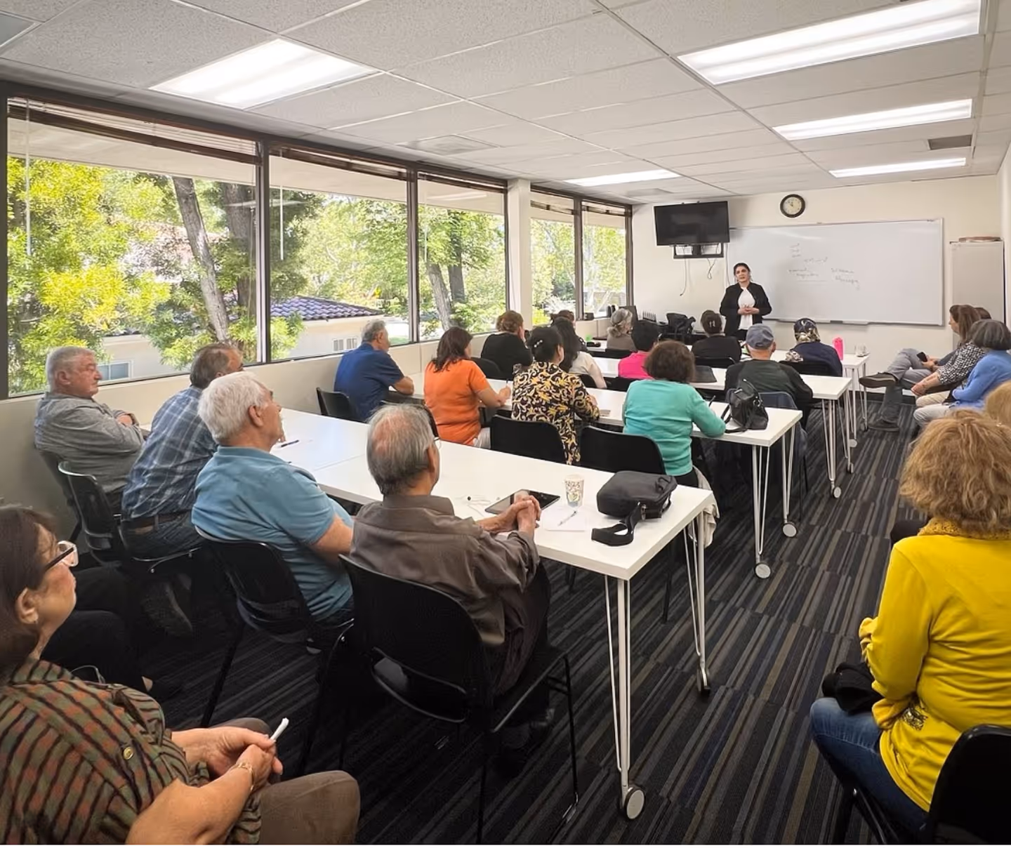 Classroom with adults seated and a teacher standing near a whiteboard at the front of the room.